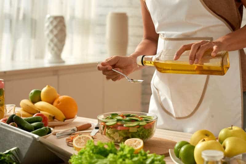 Cook wearing apron pouring olive oil into vegetable salad while standing at kitchen