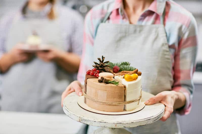 Confectioner holding tray with Christmas cake decorated with currant, cinnamon, pine cone, macaroon and kumquat