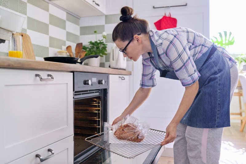 Woman puts tray of meat in the oven covered with plastic wrap