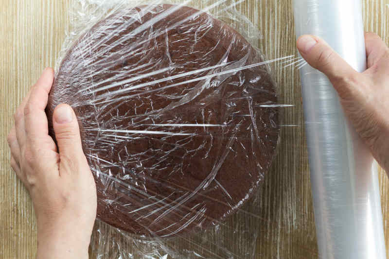 Top view of woman hand wrapping chocolate sponge cake in plastic for storing on wooden background