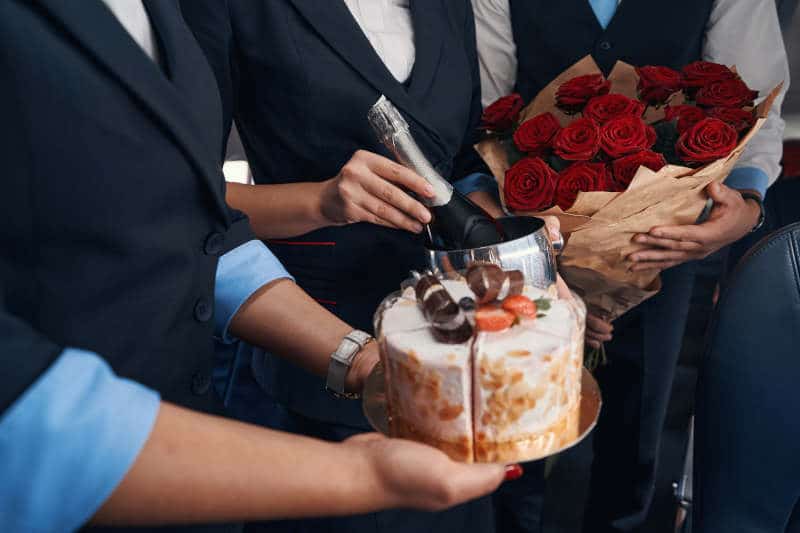People on airplane holding birthday presents for passenger, including delicious cake, champagne and roses