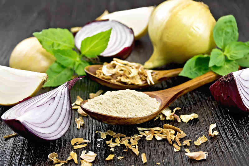 Onion powder and dried flakes in two spoons, purple and yellow onions, fresh basil on dark wooden board