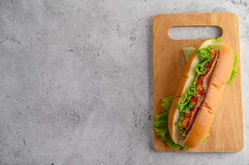 Hotdog in bread with lettuce and tomato sauce on wood cutting board