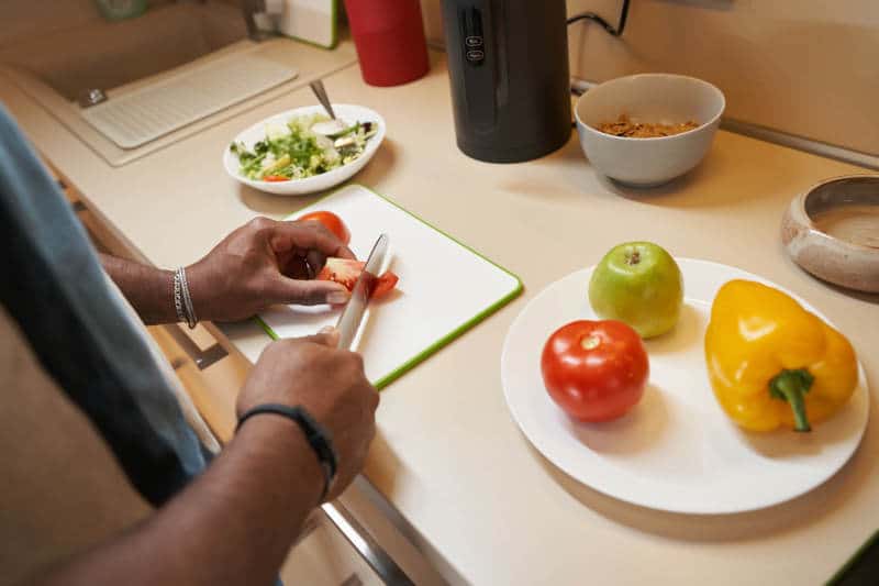 Person cutting tomato on plastic cutting board next to plate with vegetables on table