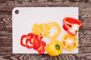 Red and yellow slices of bell pepper on white plastic chopping board over the wooden desk