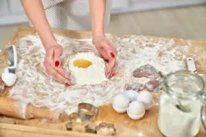 Woman making dough of baking ingredients, flour, yolk and white. Pastry board with kitchen tools as rolling pin, knife and eggs, container of flour.
