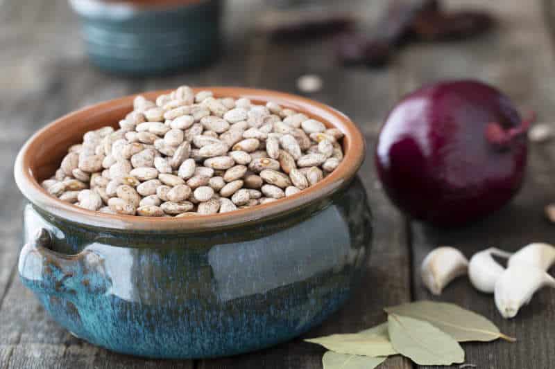 Dried pinto beans in a bowl with bay leaves, garlic and onion on old wooden table.