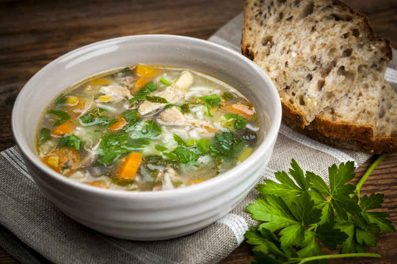 Chicken rice soup with vegetables in bowl and bread on rustic table