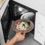Woman putting plate of rice with vegetables in microwave oven