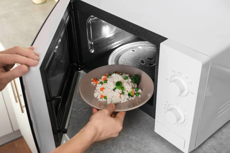 Woman putting plate of rice with vegetables in microwave oven